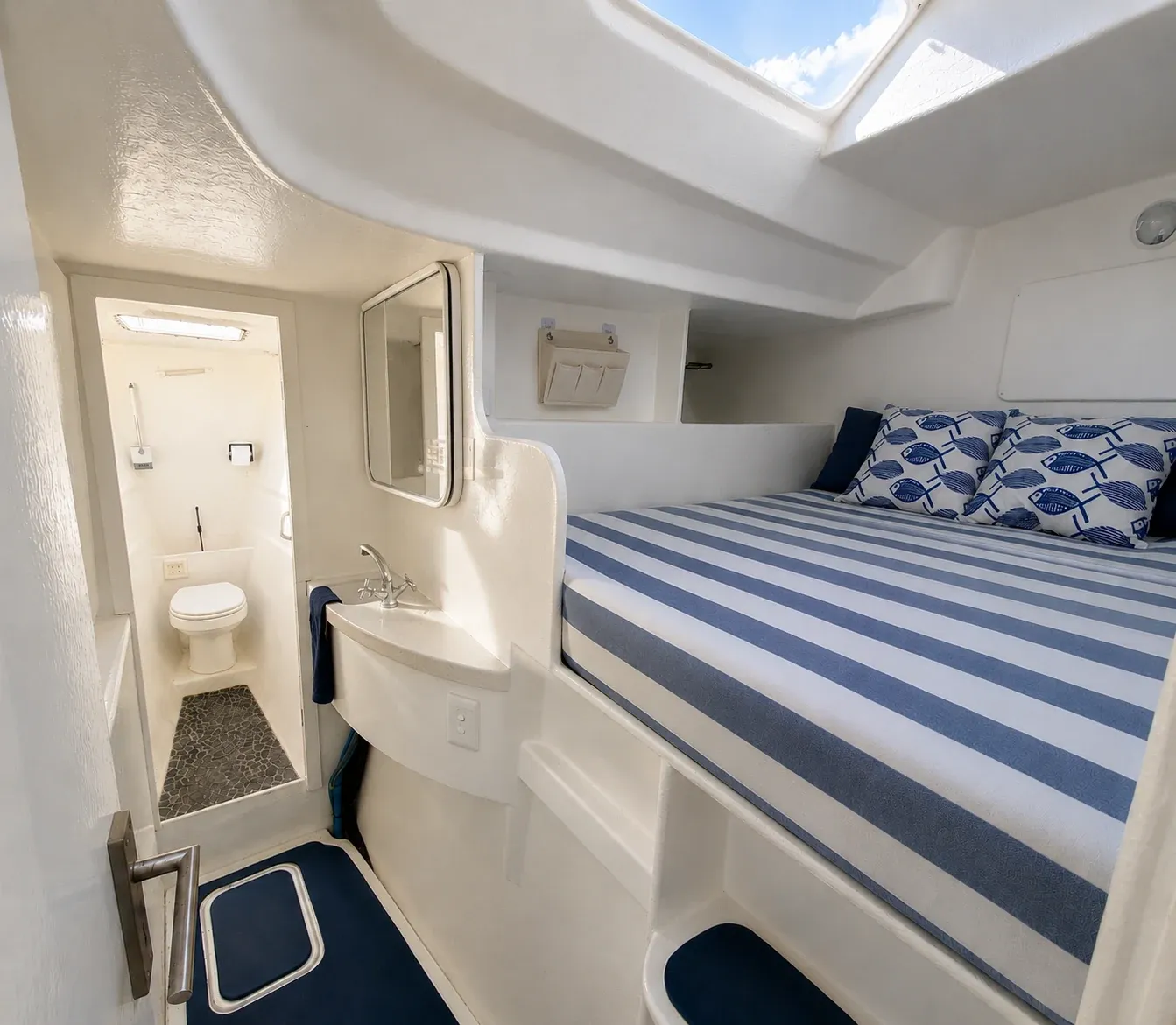 Bright forward double cabin with a striped berth, nautical scatter cushions, an overhead hatch, and an ensuite to one side.