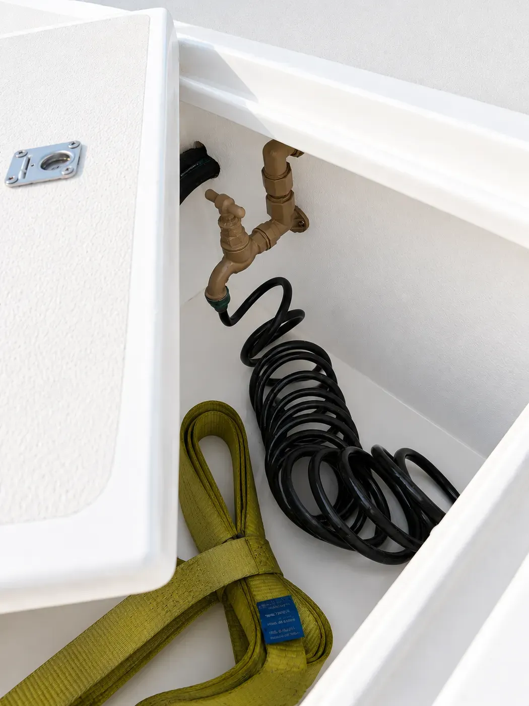 Open foredeck wet locker aboard Vaquita with a brass washdown tap, coiled high-pressure seawater hose, and a lifting strap.