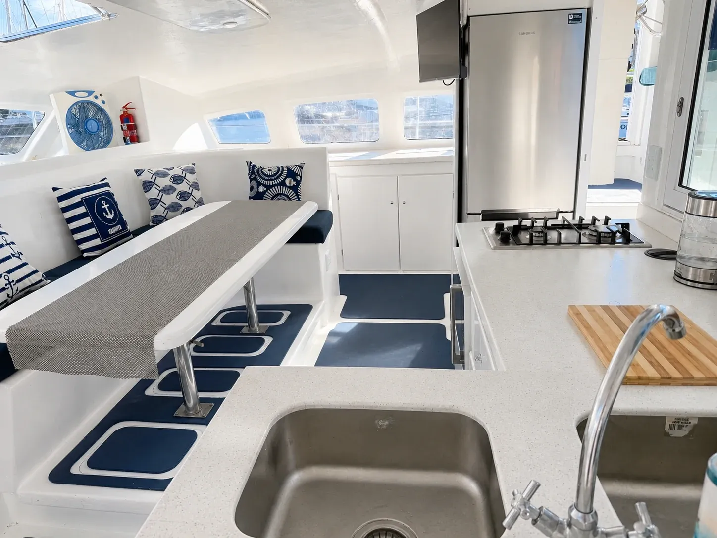 Saloon looking through to the galley aboard Vaquita, with bench seating, gas hob, double sink, and stainless fridge.