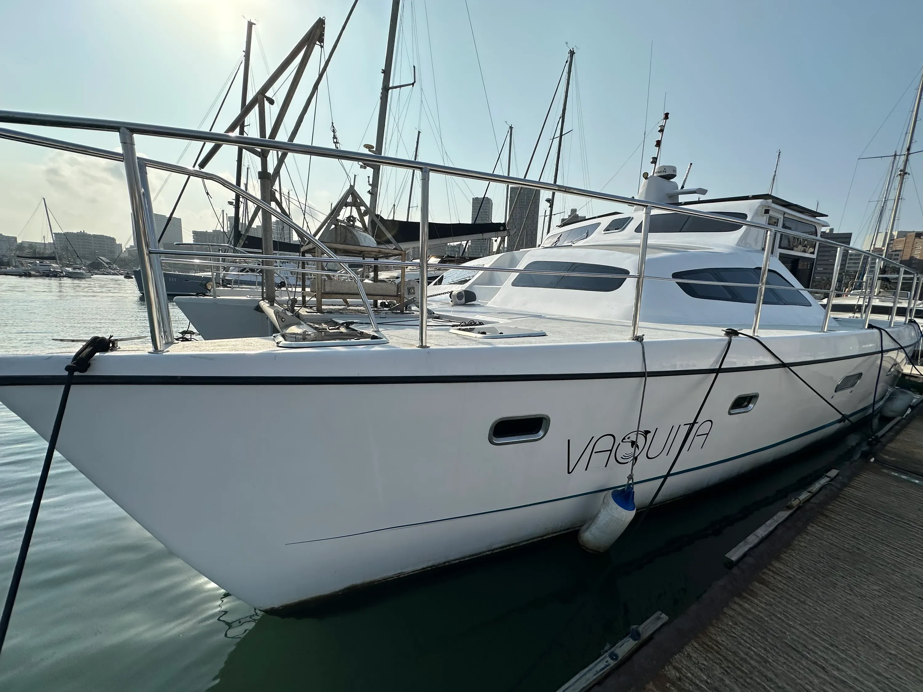Vaquita moored bow-on at her Durban Harbour berth, port-bow view showing the white catamaran hulls, the wheelhouse, and the masts of the marina behind.