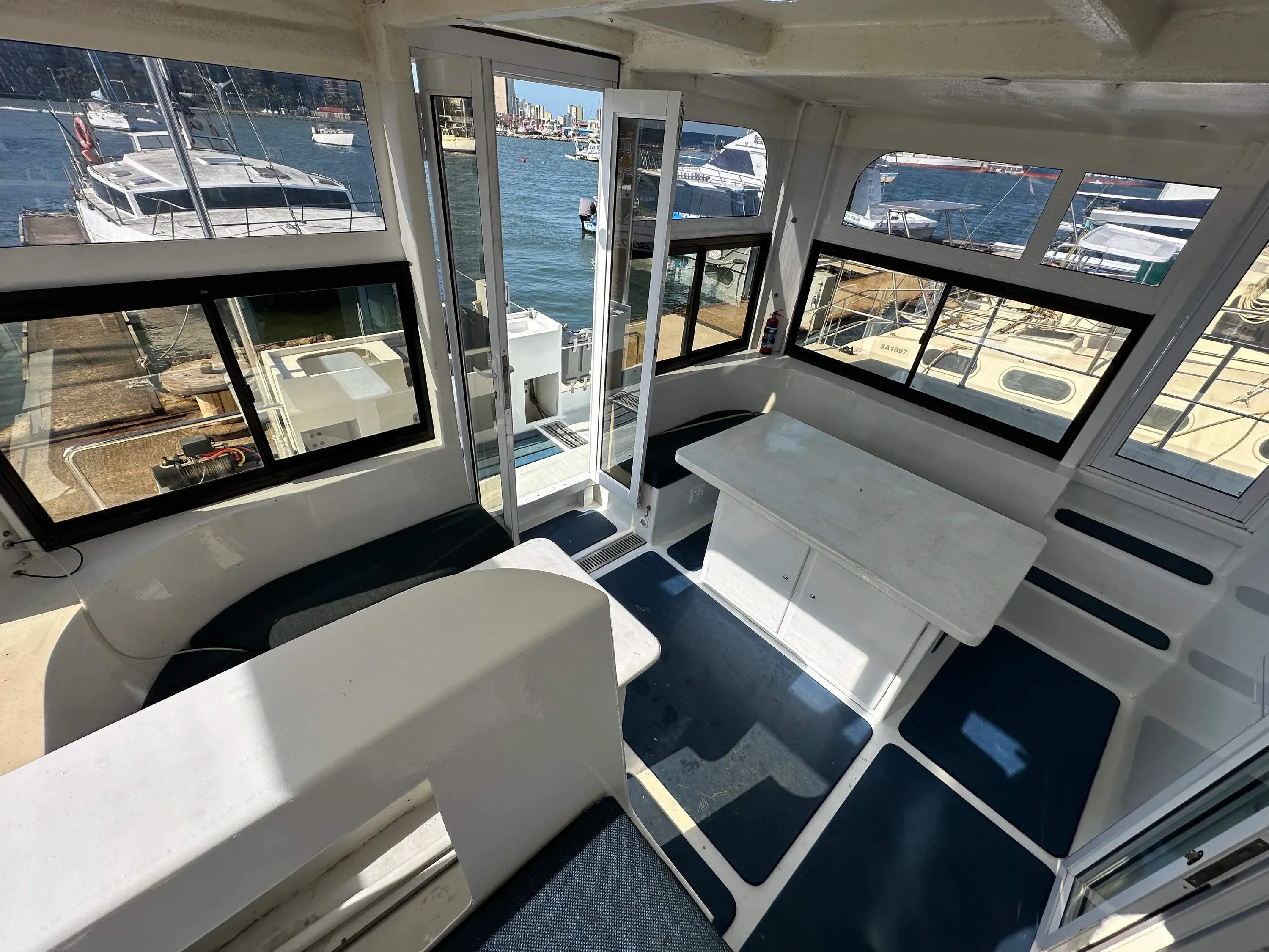 Inside Vaquita's wheelhouse looking out through the saloon windows, with the chart table, bench seating, and the marina visible beyond.