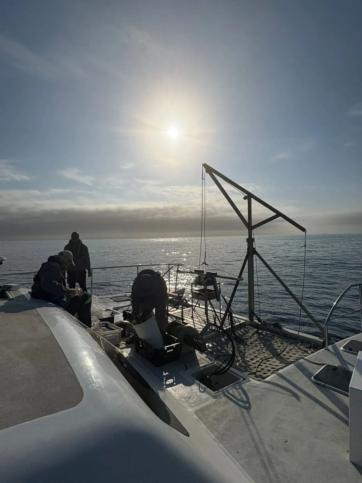 Crew working Vaquita's foredeck at first light, the A-frame davit rigged and the low sun reflecting off a calm sea.