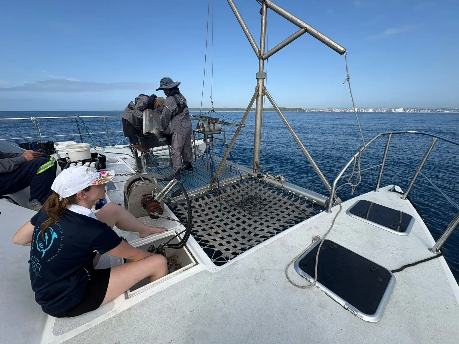 Crew working on Vaquita's foredeck offshore, one seated taking notes in a Kunye Sea polo, with the trampoline net and coastline in the distance.