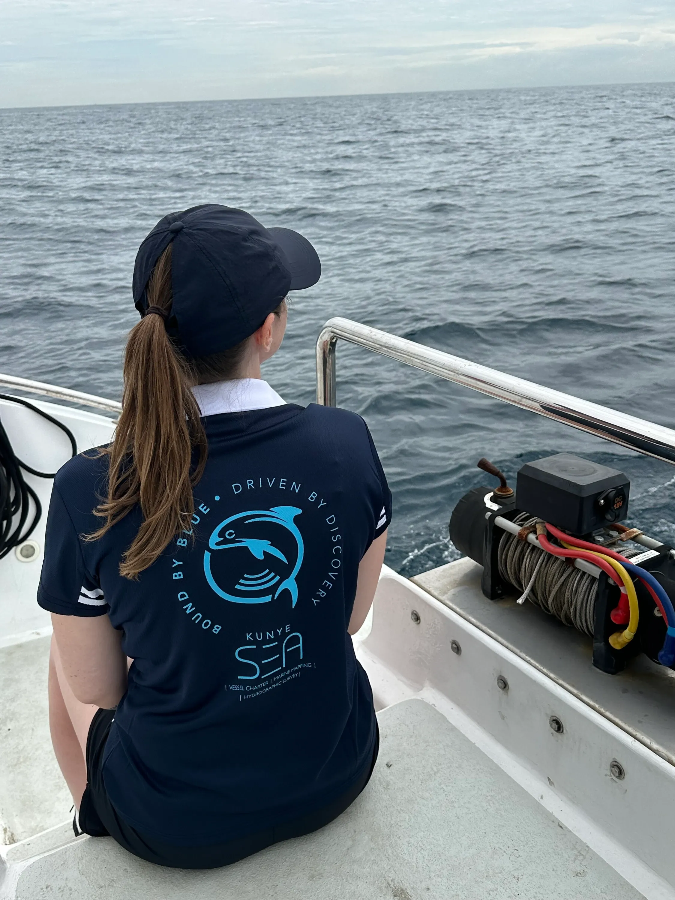 Crew member in a Kunye Sea polo, back to camera, looking out to sea from Vaquita's quarter. The 'Bound by Blue. Driven by Discovery.' tagline arcs around the dolphin brand mark.