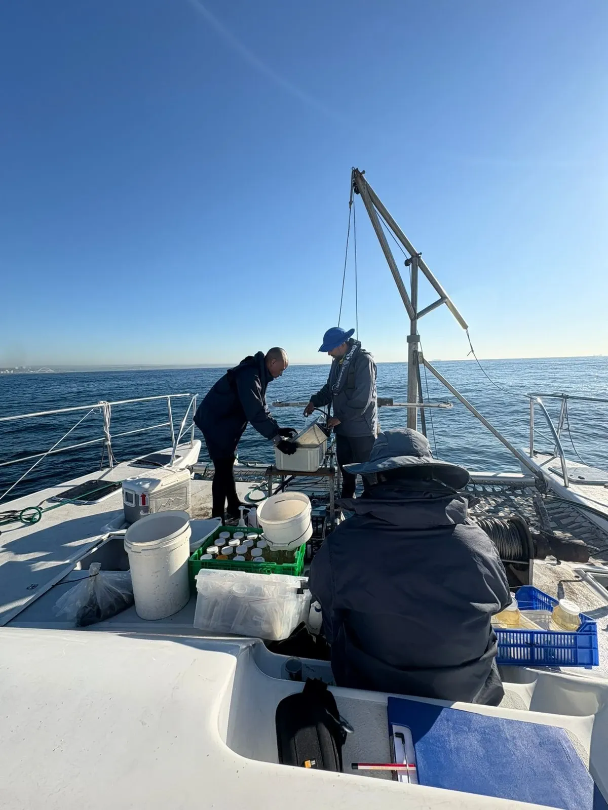 Crew sampling on Vaquita's foredeck offshore, working over a sediment sampler with sample bottles and buckets crated on deck under a blue sky.