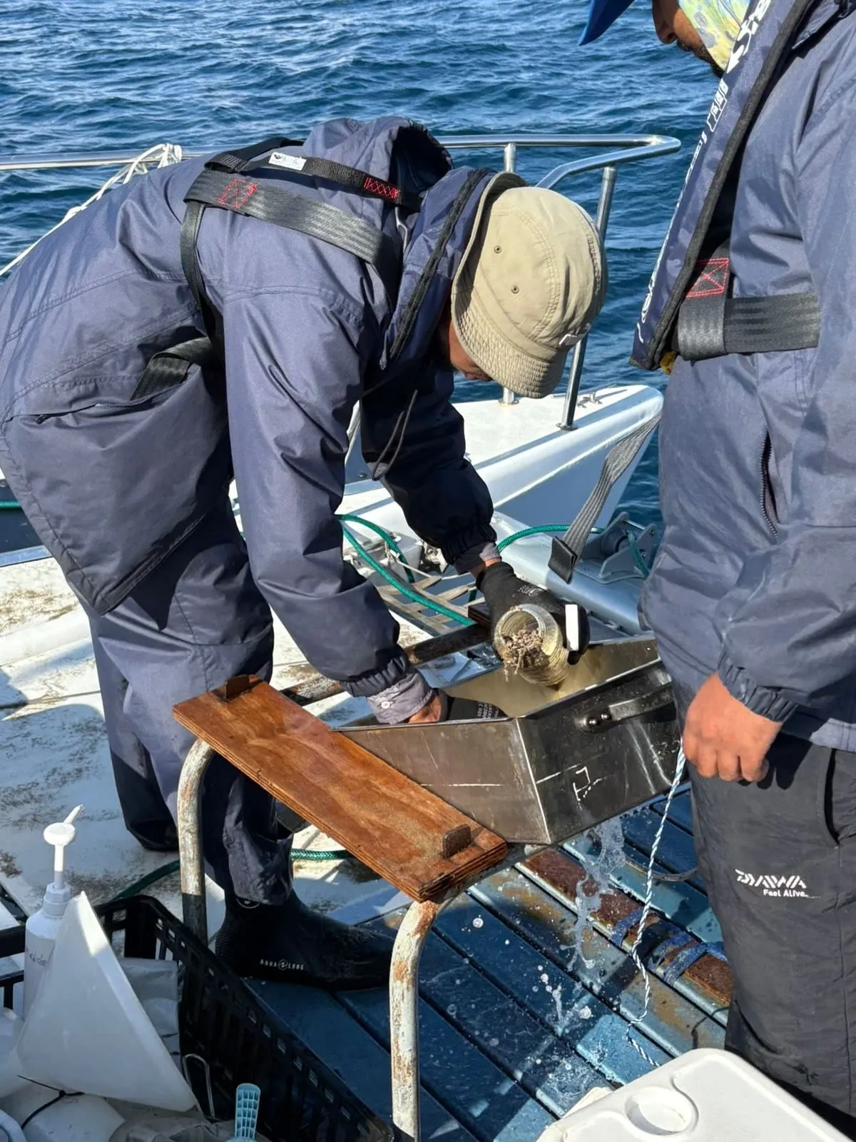 Two crew in foul-weather gear emptying a stainless grab sampler onto a sorting board on Vaquita's stern, seawater draining as they work.