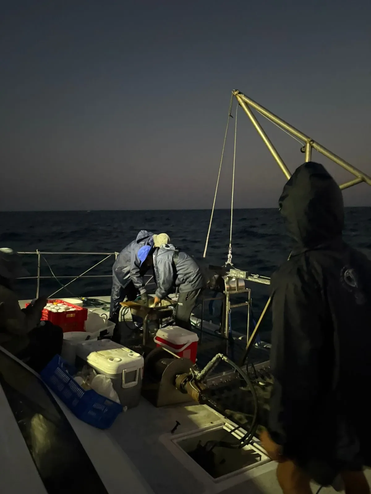 Crew working Vaquita's foredeck at dusk under the A-frame davit, cooler boxes and gear crates on deck and a calm sea fading into twilight.