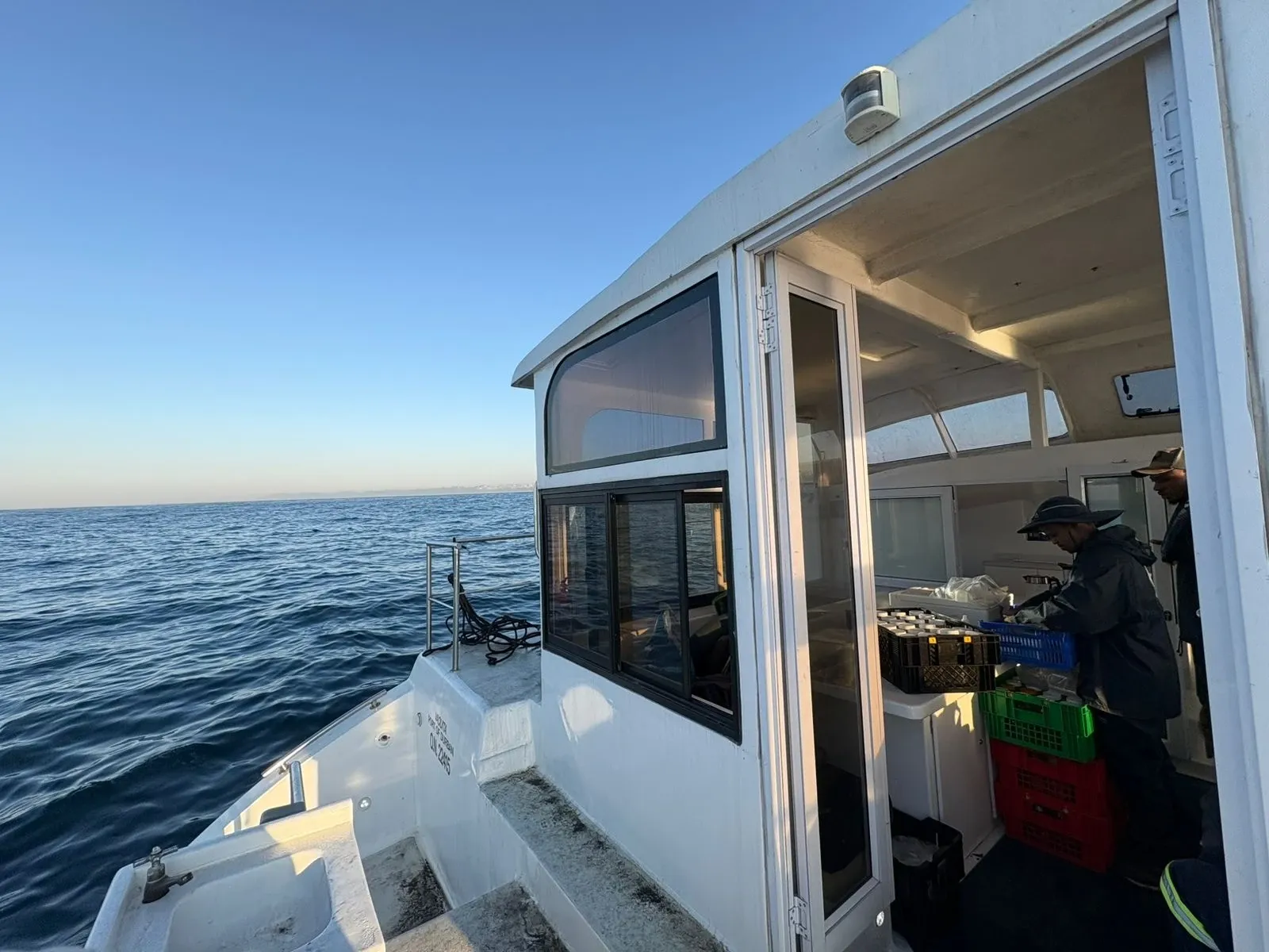 Looking aft along Vaquita's deck at golden hour toward the open wheelhouse door, where a crew member sorts and categorises sample bottles into crates, the calm sea alongside.