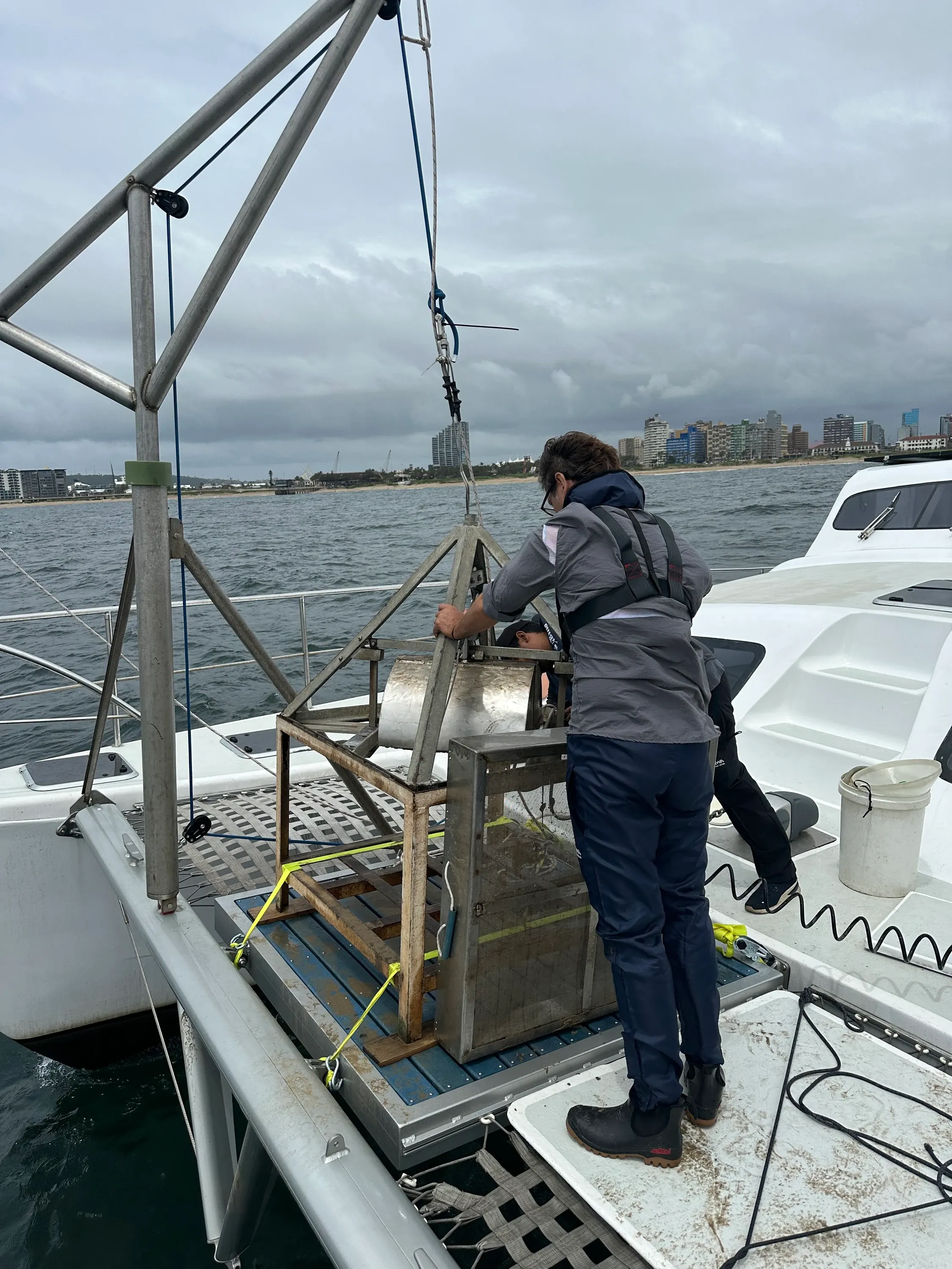 Crew member operating the stainless grab sampler on Vaquita's A-frame davit at the stern scoop, the Durban city skyline on the horizon behind.